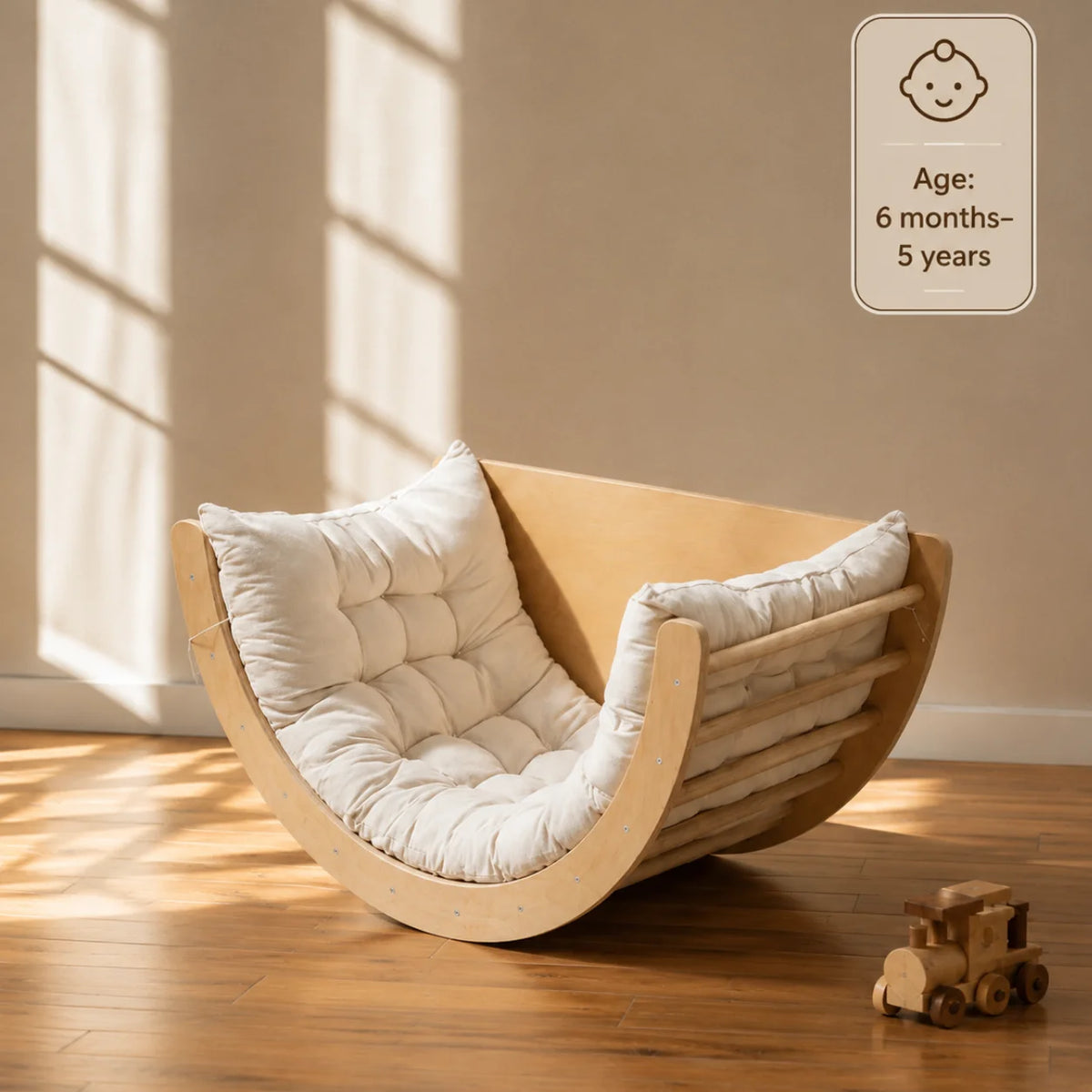 A curved wooden baby floor chair with thick white tufted cushions, placed on a wooden floor in a sunlit room. A small wooden toy train sits nearby, and a badge in the top right reads 'Age: 6 months-5 years'.