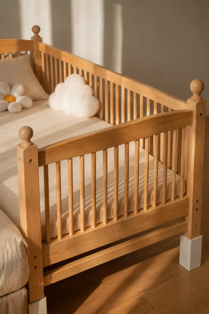 A light natural wood toddler bed with safety slatted sides, styled with a white mattress, cream bedding, and a white cloud-shaped pillow in a softly lit room.