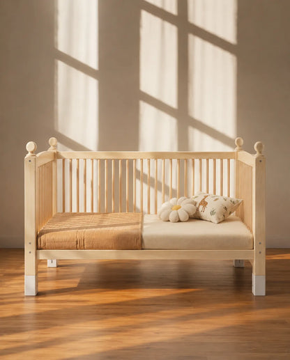 A light natural wood baby crib with white-painted legs, styled with a tan blanket and a cream flower-shaped pillow on a wooden floor, set against a wall with warm window shadow patterns.