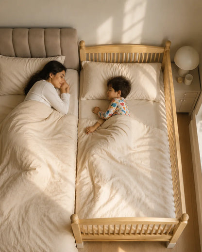 A woman and a young child rest under cream bedding in two adjacent beds, one full-size bed and one wooden toddler bed, in a softly lit bedroom.
