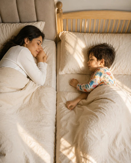 A woman and a young child sleep side by side in separate beds with matching cream bedding, lit by warm natural sunlight.