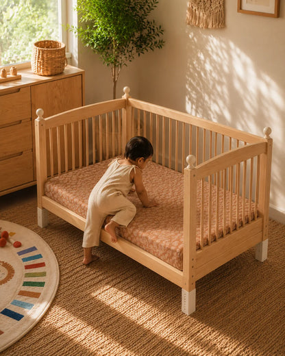 A baby in a cream outfit crawls on a patterned mattress inside a light natural wood toddler bed with white feet, set in a warm, sunlit nursery.