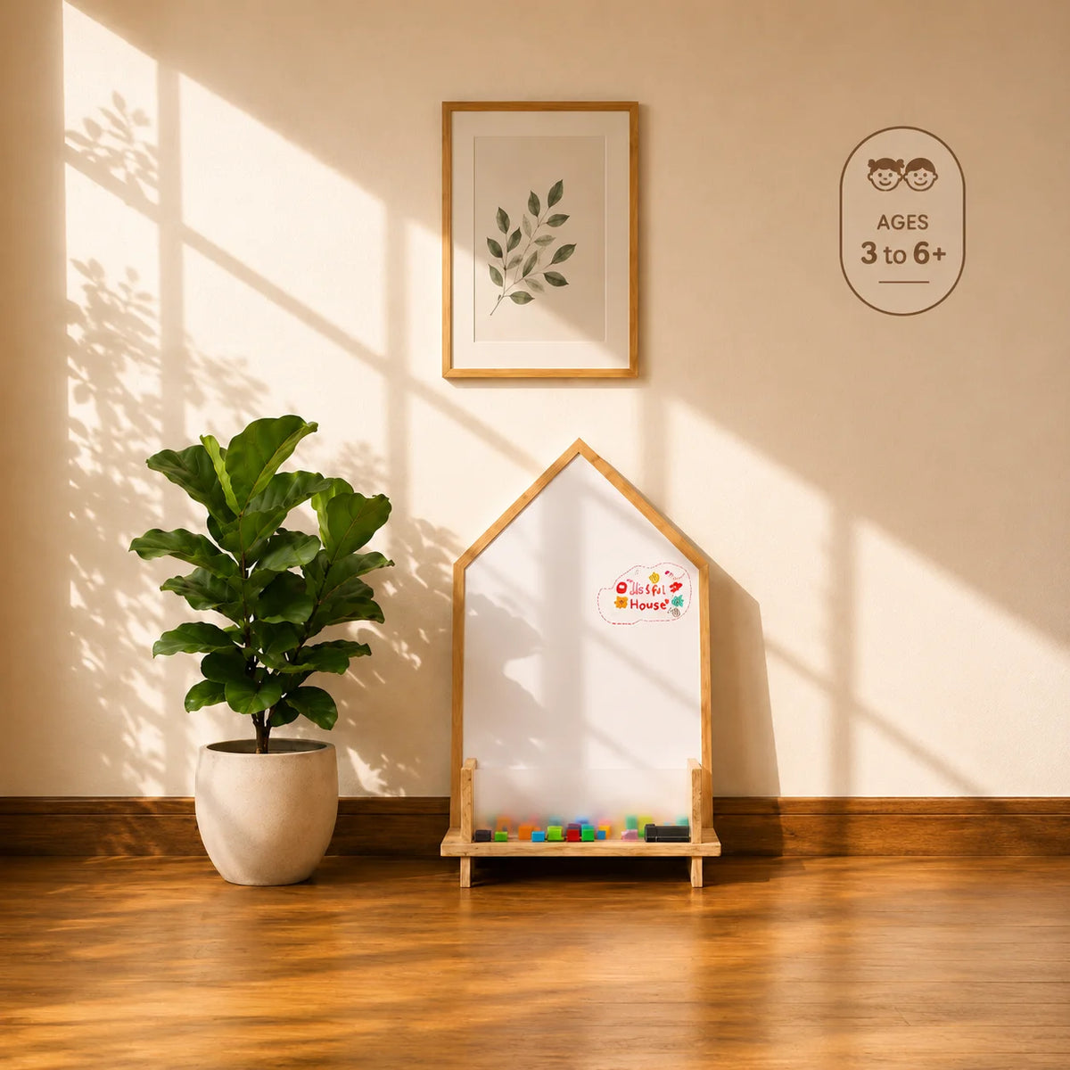 A house-shaped whiteboard with a light wood frame stands on a wooden floor against a beige wall, with warm sunlight casting shadows across the scene. A potted green plant is on the left, and a framed leaf print hangs on the wall above.