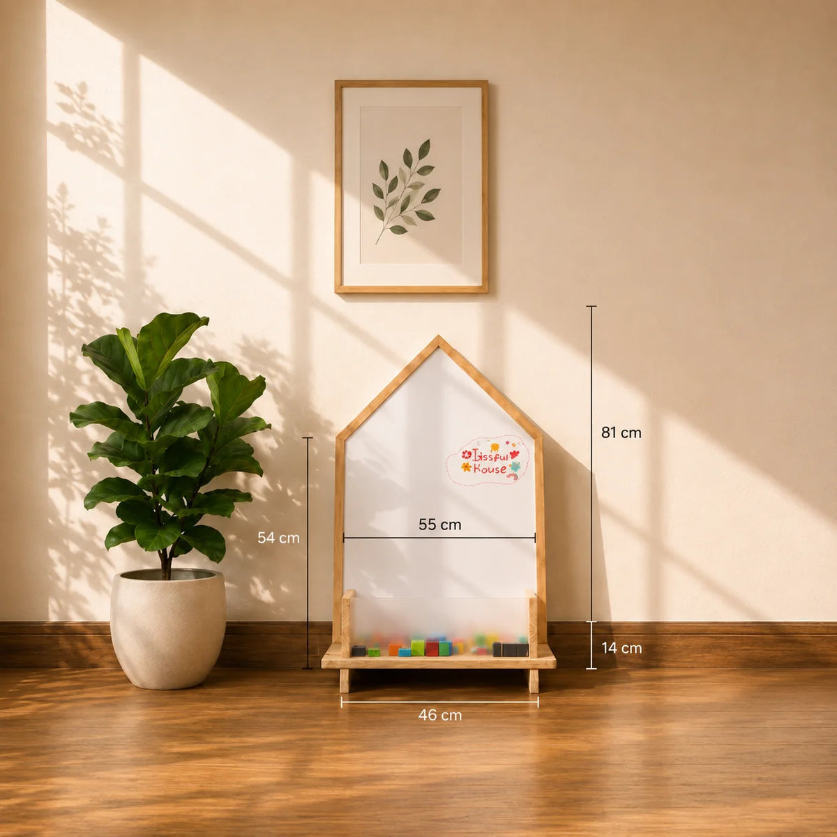 A house-shaped children's activity board with a white writing surface, wooden frame, and colorful blocks in the base, shown in a styled room with wall art, a potted plant, and dimension labels overlaid on the image.