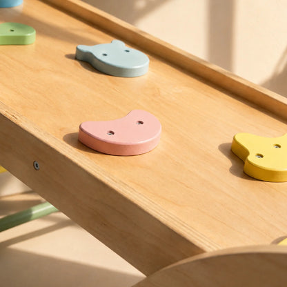 Close-up of a wooden children's climbing or sliding toy with pastel animal-shaped hand grips in pink, light blue, yellow, and green on a natural wood surface.