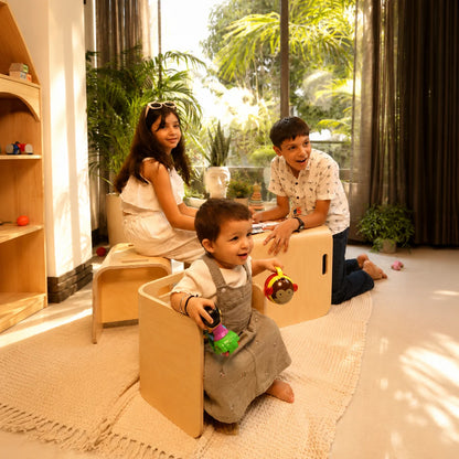 A family of three in a bright, modern playroom with wooden children's furniture. A young child sits in a light wood chair in the foreground, while a woman and a man relax on matching wooden chairs and a bench in the background.