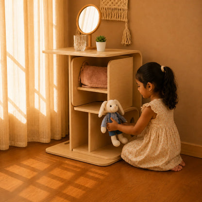 A young girl in a beige dress kneels on a wooden floor next to a light wood storage cabinet, holding a white bunny plush toy. The cabinet has open shelves, one holding a pink bag, and the top shelf has a small mirror and a plant.