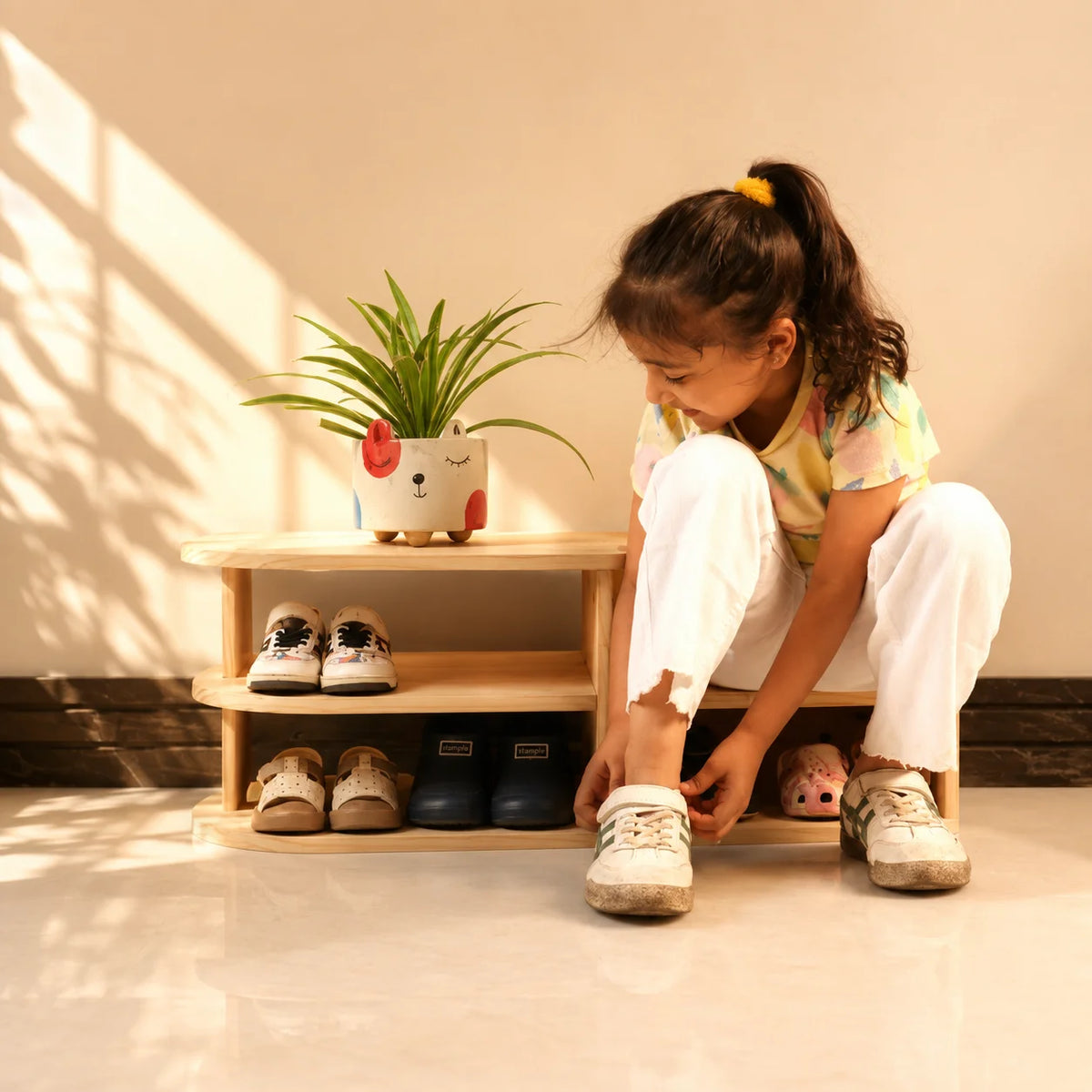 A young girl sits on a small wooden shoe rack in a brightly lit room, tying the laces of her white sneakers. The rack holds several pairs of shoes and a potted plant on top.