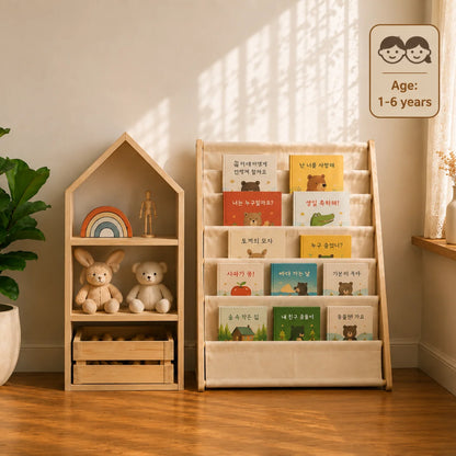 A styled children's room corner with a wooden house-shaped bookshelf and a fabric book rack filled with children's books, placed on a woven rug beside a bed and window.