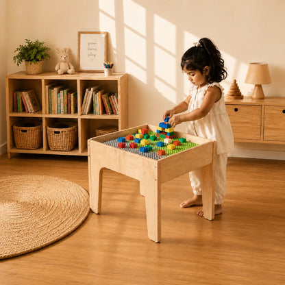 A young girl in a white dress stands barefoot next to a light wood activity table, stacking colorful building blocks on a green grid. The scene is set in a warm, sunlit room with wooden floors, a bookshelf, and a woven rug.
