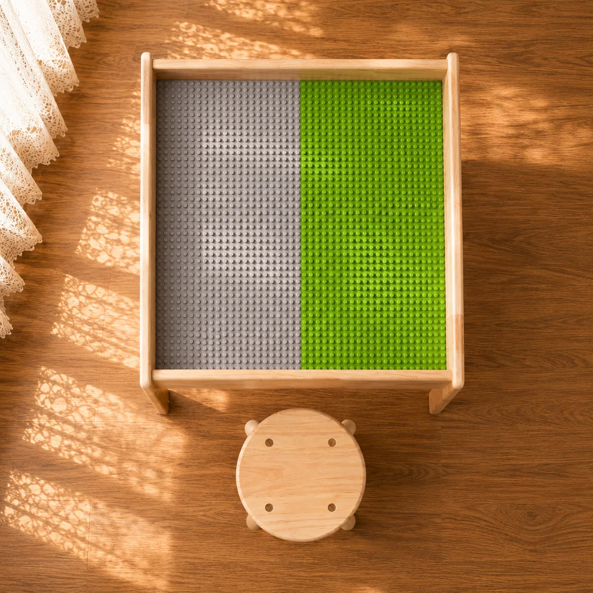 A square wooden children's table with a textured insert split vertically into gray and green sections, shown from above on a wooden floor with a small round wooden stool below it.