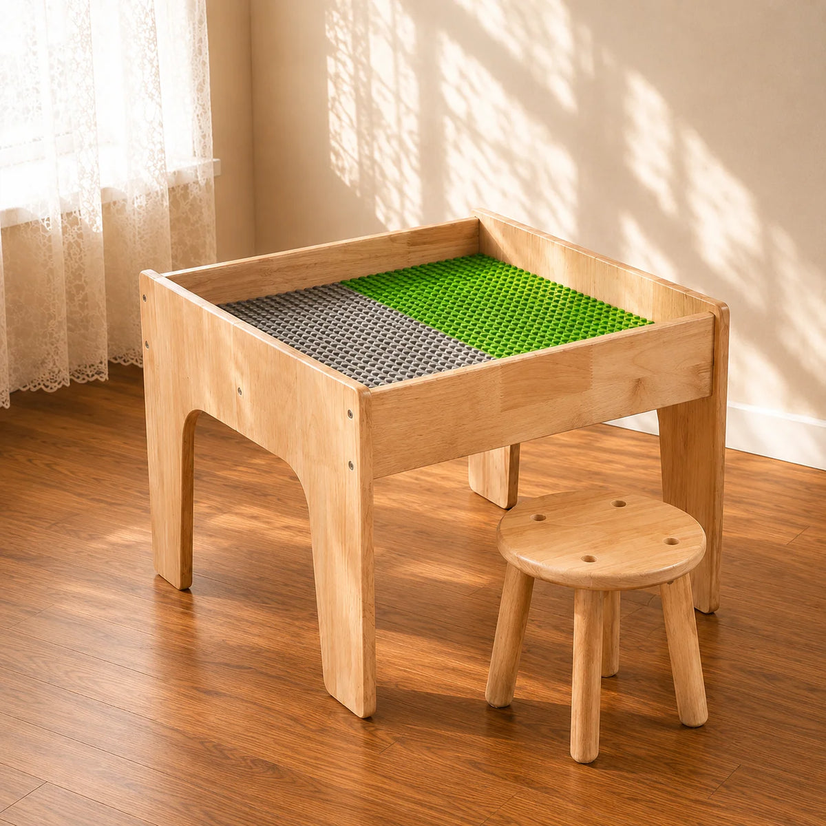 A light natural wood children's activity table with a green and gray textured play mat inside, shown with a matching small wooden stool in a sunlit room with a wood floor and cream walls.