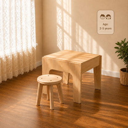 A light natural wood children's table with a matching round wooden stool in a sunlit room with warm beige walls, lace curtains, and a potted plant. A small sign on the wall reads 'Age: 2-5 years'.