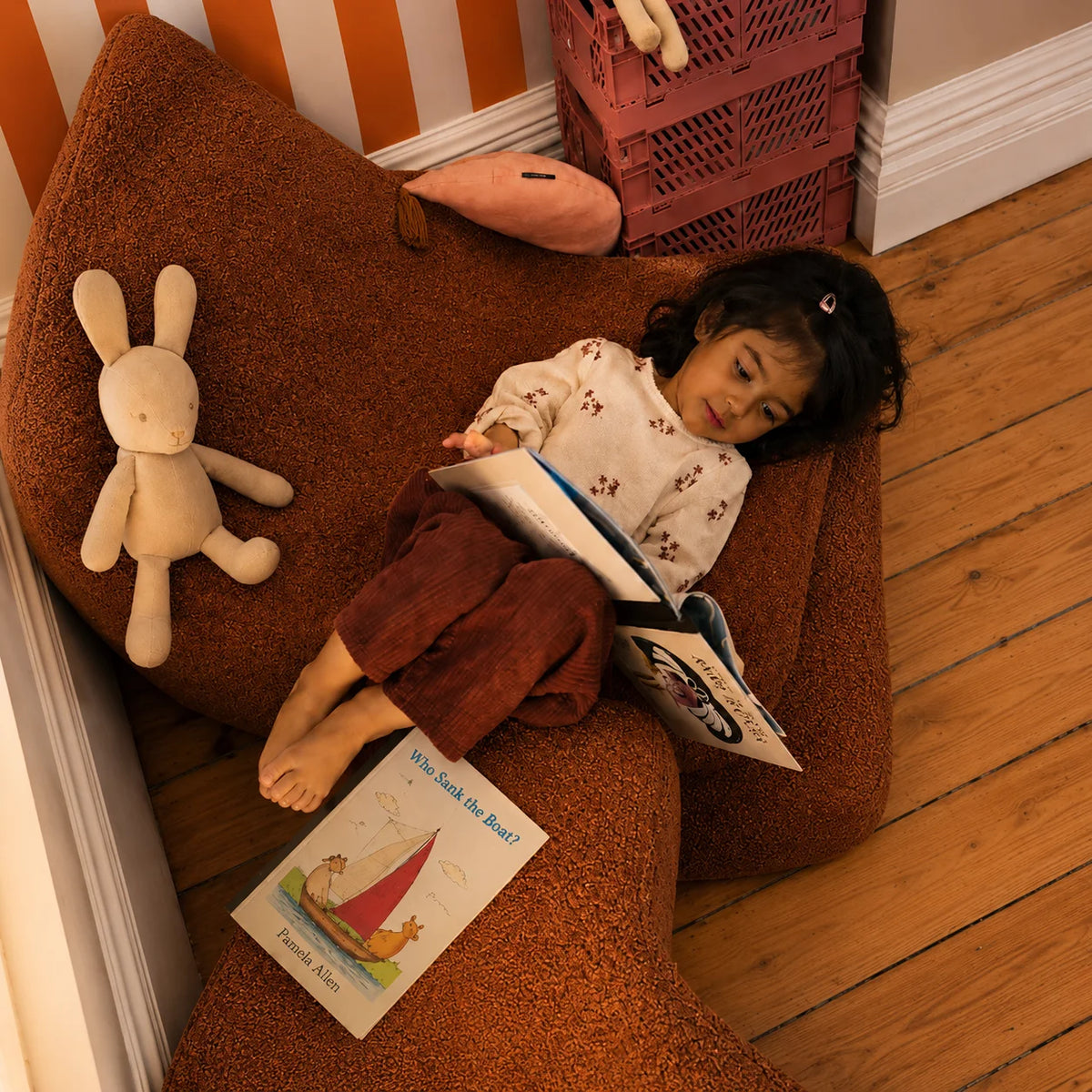 A young child in a light pink dress stands in front of a wooden toy and book storage shelf in a warm, sunlit room. The shelf contains colorful children's books, plush toys, and small decorative items.