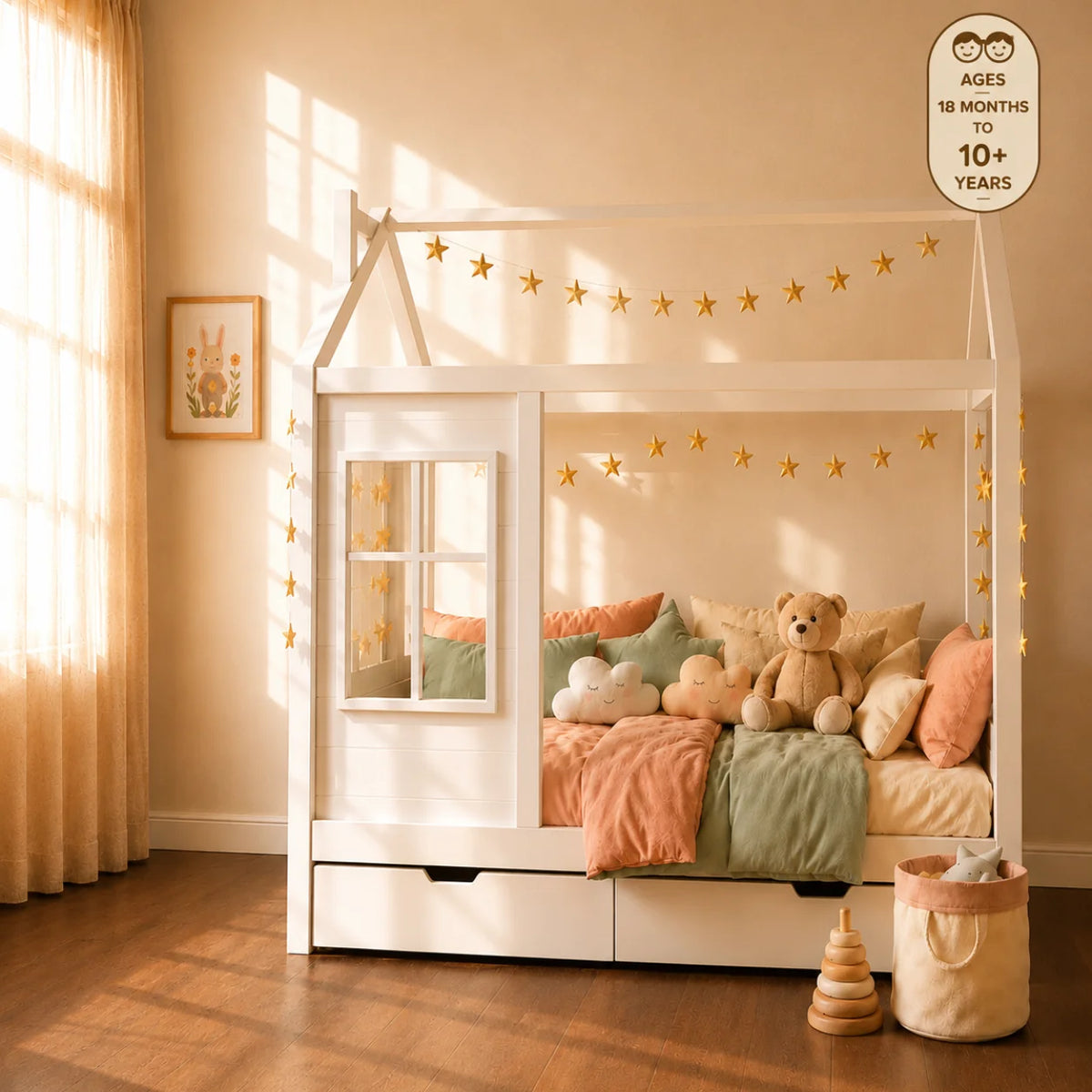 A white house-frame toddler bed with hanging star and ball garlands, shown in a warm, sunlit bedroom with a man and woman sitting on the bed and a young girl playing beside it.