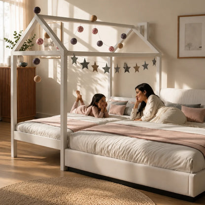 A white house-shaped canopy bed in a sunlit child's bedroom, with a woman and a young child sitting inside on a quilted bed. The canopy is decorated with hanging metallic ornaments and a star garland.