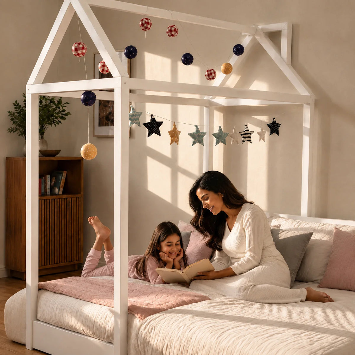 A white house-frame canopy bed in a warm, sunlit bedroom, with a woman and child resting on the bed with pink bedding and pillows.