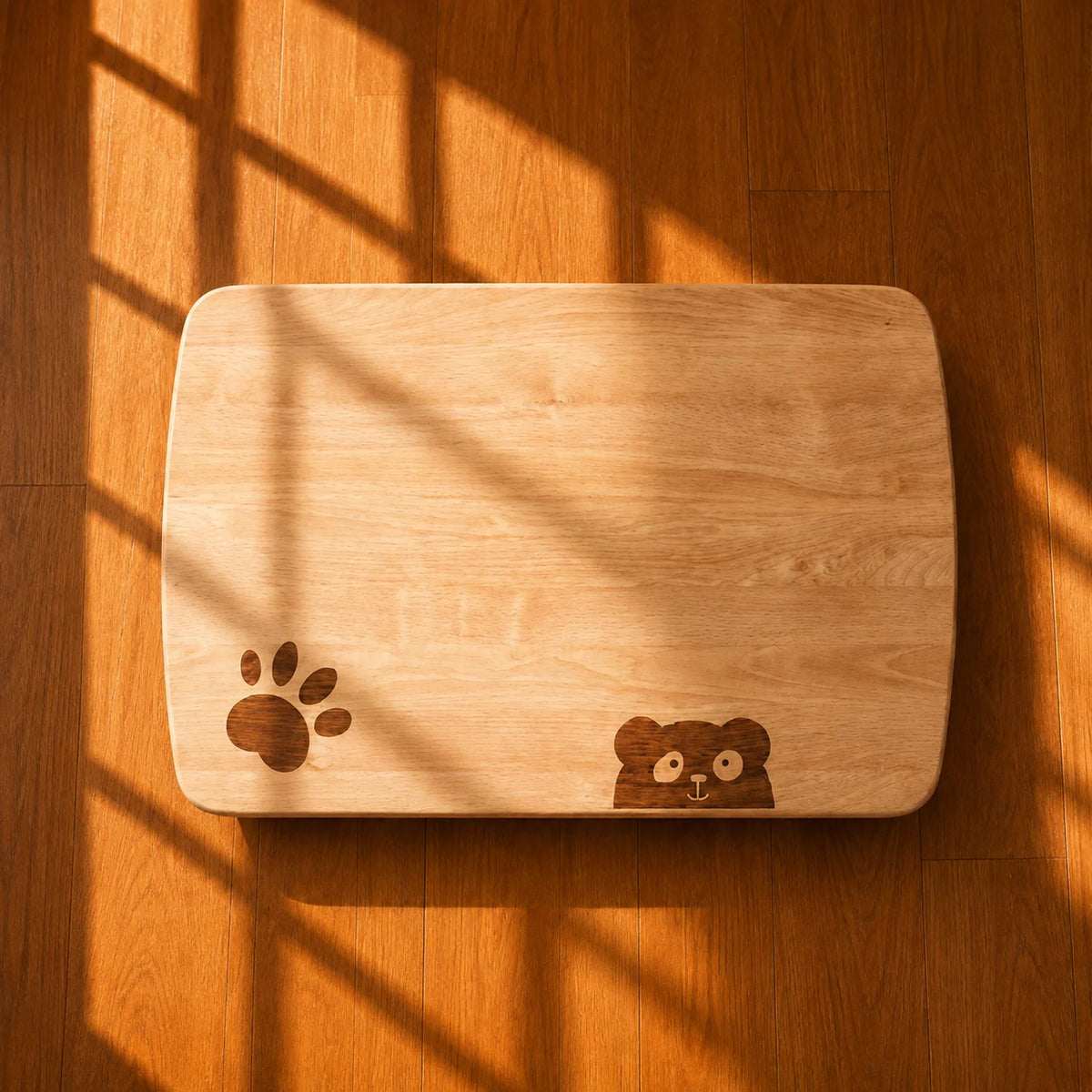 A rectangular wooden cutting board with rounded corners, featuring a dark brown paw print engraving on the bottom left and a small bear face engraving on the bottom right. The board is photographed from above on a warm wood floor with strong window shadow patterns.