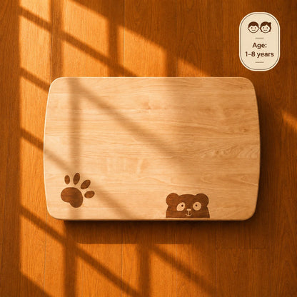 A rectangular wooden cutting board with rounded corners, featuring a dark brown paw print and a small bear face engraving, displayed on a warm wood floor with dramatic window shadow patterns.