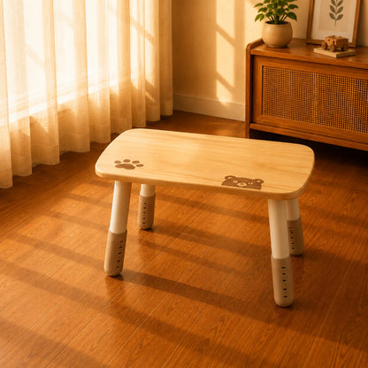 A small wooden children's table with rounded corners, a light natural wood top, and white legs with gray footrests, placed on a warm wood floor in a sunlit room near a window and cabinet.