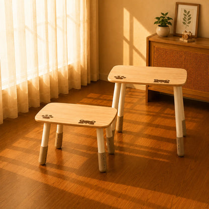 Two small wooden stools with white legs and paw-print details, placed on a warm wood floor in a sunlit room with curtains and a cabinet in the background.