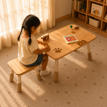 A young girl sits at a small wooden children's table with a matching bench in a warm, sunlit room. The table has a light natural wood top with darker brown printed paw prints and legs, and is styled with toys and a book.