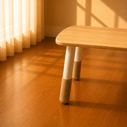 A modern wooden low table with tapered legs, shown in a warm, sunlit room with a wood floor and sheer curtains.