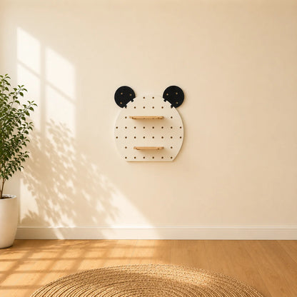A wall-mounted wooden shelf unit with two black circular pegs at the top resembling ears, mounted on a beige wall in a sunlit room with a plant and wood floor visible.