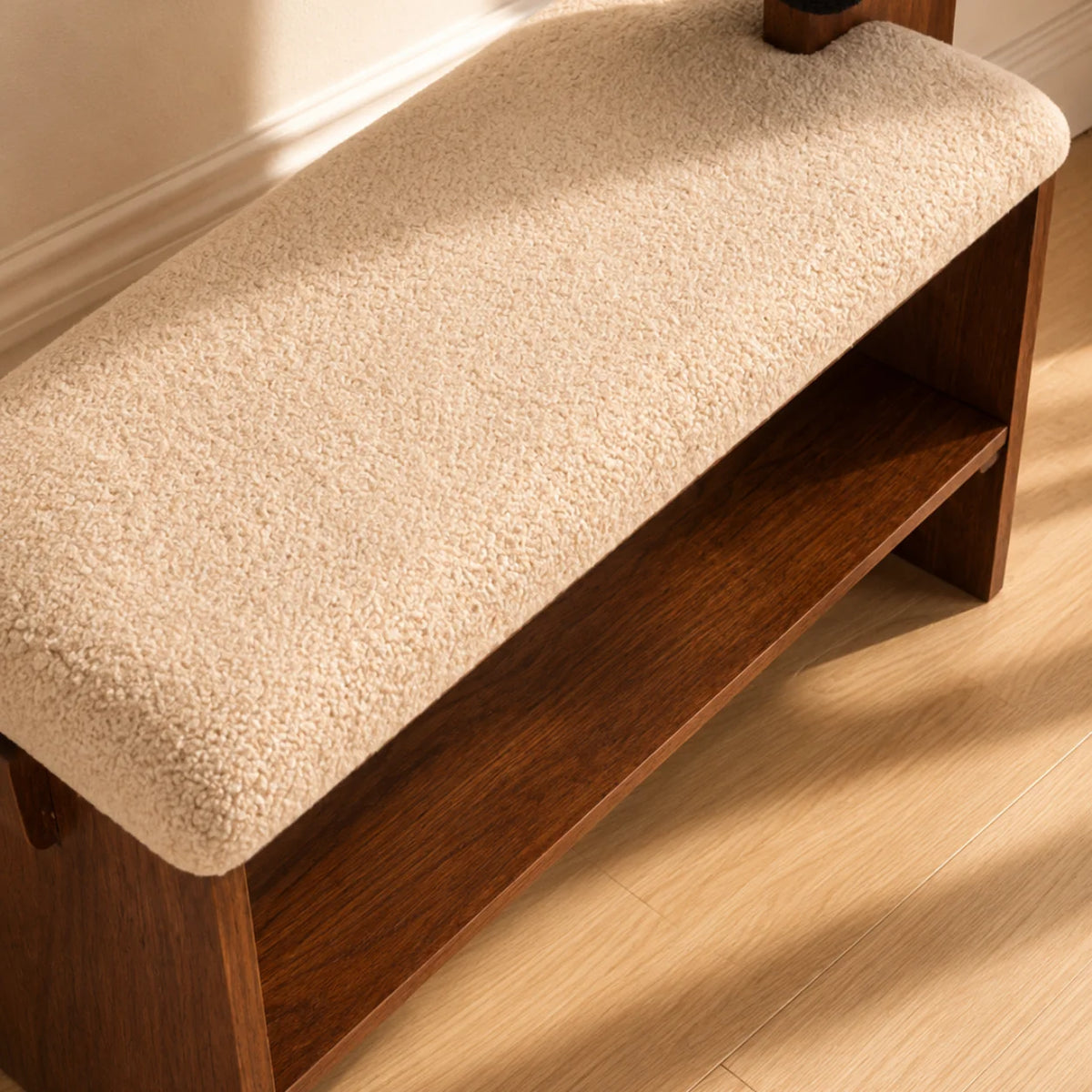 A wooden entryway bench with a thick, textured cream bouclé cushion on the seat and a lower shelf, shown on a light wood floor in warm sunlight.