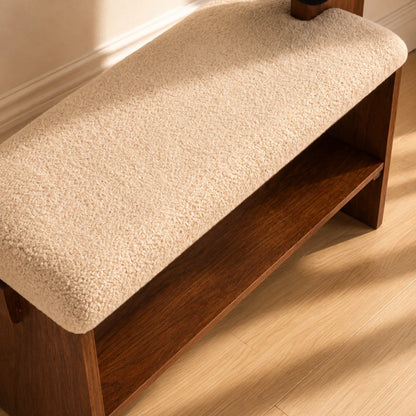 A wooden entryway bench with a thick, textured cream bouclé cushion on the seat and a lower shelf, shown on a light wood floor in warm sunlight.