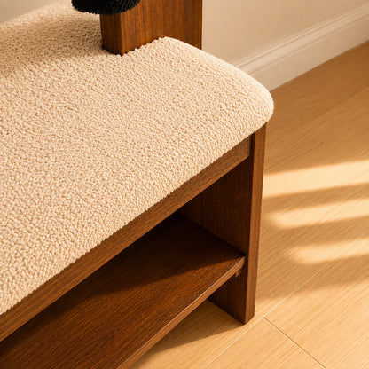 A close-up of a wooden bench or shelf with a thick, textured cream boucle cushion on the seat, set on a light wood floor with warm sunlight casting shadows.