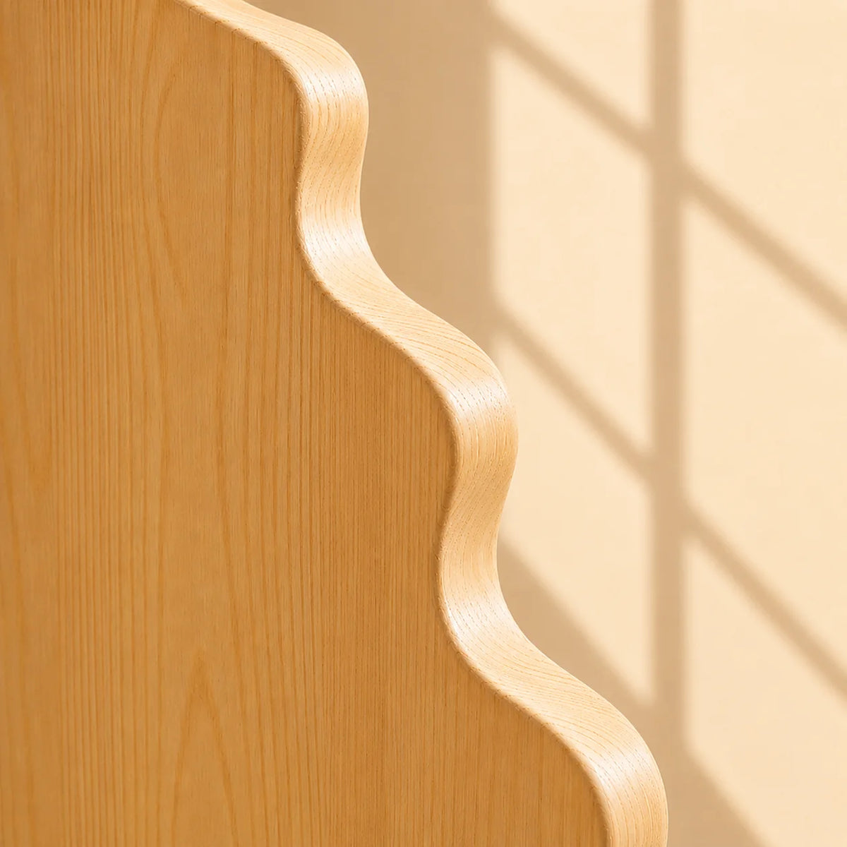 Close-up of a light natural wood panel with a wavy scalloped edge against a warm beige background with soft window-shadow lines.