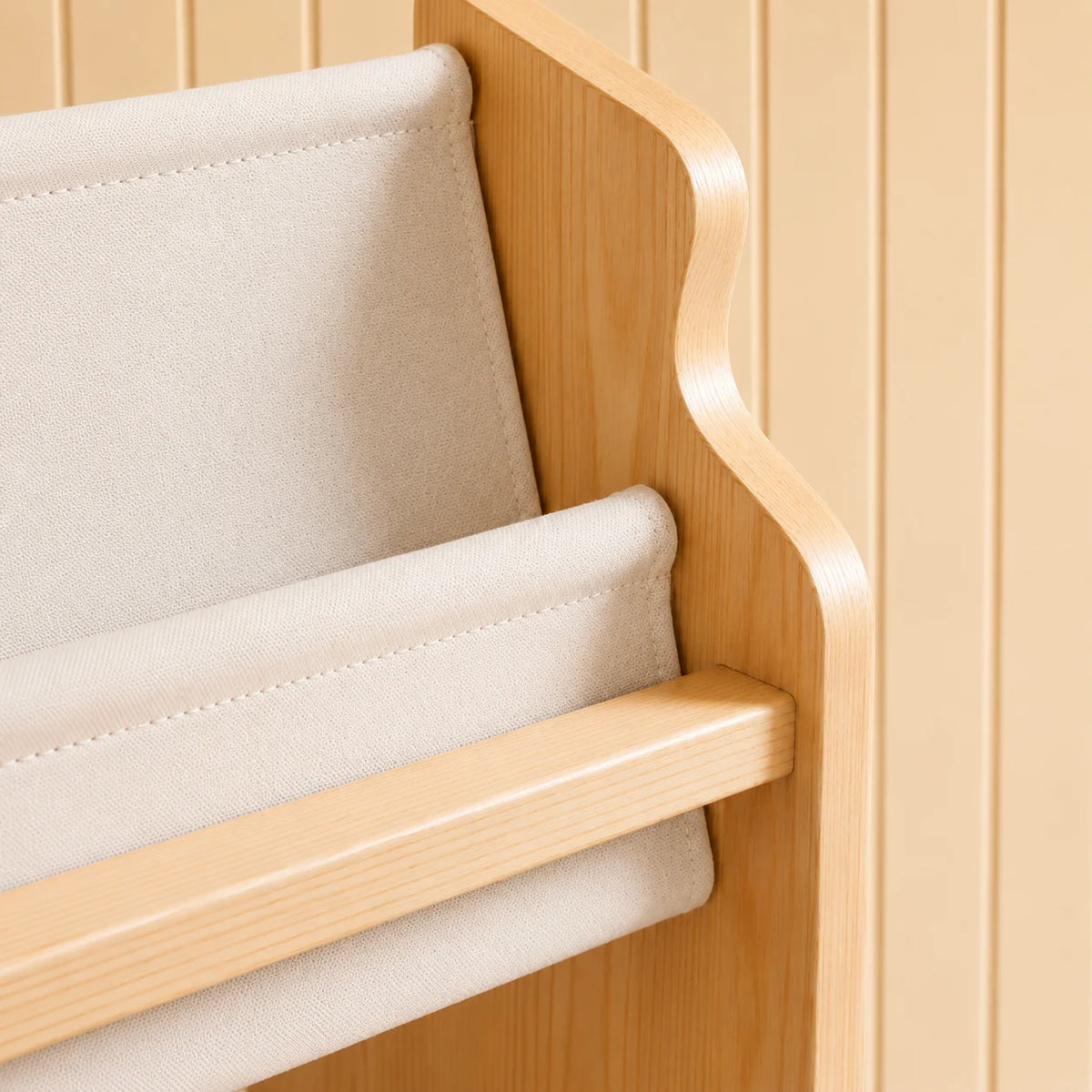 Close-up of a light natural wood bookshelf or bookcase with cream fabric shelves, set against a matching beige wood-paneled background.