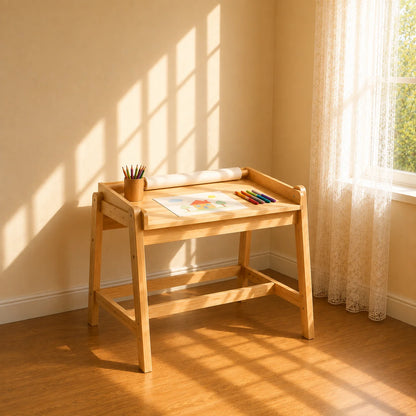 A young child stands barefoot at a small wooden writing desk in a sunlit room, drawing on paper. The room has warm wood floors, sheer curtains, framed animal artwork on the wall, and a potted plant by the window.
