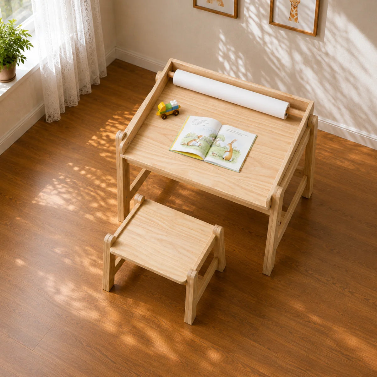 A light natural wood children's writing desk with an attached matching stool, shown in a sunlit room with hardwood floors, a window, curtains, framed wall art, and a few small desk props including an open book, a toy car, and a rolled sheet.